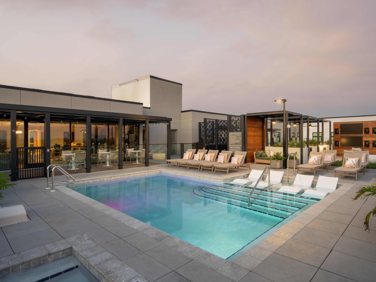 A modern rooftop pool area at sunset features lounge chairs, in-water loungers, potted plants, and glass walls surrounding the Sweetwater pool. Relaxing seating areas and a stylish, open indoor lounge are visible in the background.