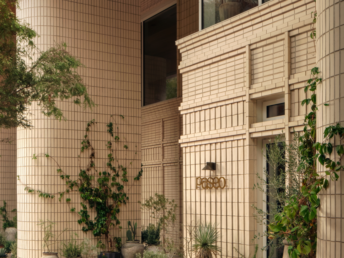 A modern building with cream-colored rectangular tiles, large windows, and potted plants lining the base creates a welcoming paseo. Various green leafy plants and small trees thrive while reddish bricks cover the ground.