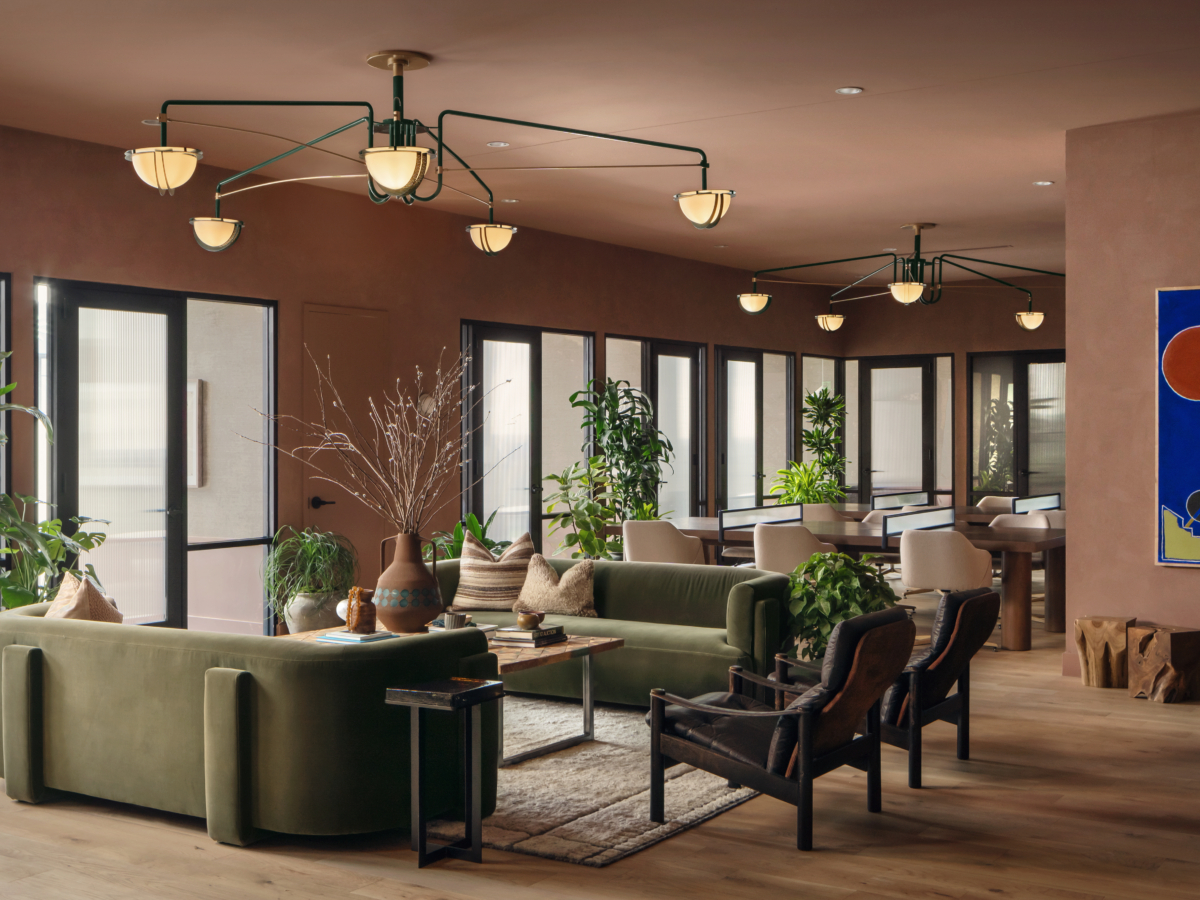 A modern lounge at Paseo with green sofas, black armchairs, large windows, and indoor plants. Overhead, unique chandeliers hang from a brown ceiling. A blue abstract painting decorates one wall, and wood flooring adds warmth.