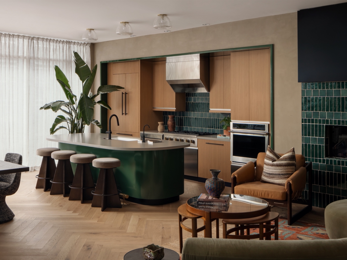 Modern kitchen and living area with wood cabinets, green tile accents, an island with dark stools, plants, a fireplace, and cozy seating. Natural light filters through sheer curtains along the stylish Paseo, highlighting sophisticated decor.