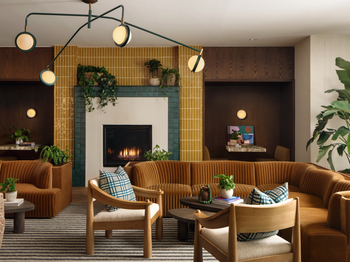 A stylish lounge at Paseo with curved brown sofas, wooden chairs, a lit fireplace framed by yellow tiles, green plants, round light fixtures, and cozy alcoves with tables against a striped rug and neutral walls.