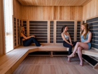 Three women sit and chat together in a modern wooden Sweetwater sauna, two on the right and one on the left, all relaxed and casually dressed in fitness wear. Sunlight enters from a window on the left.