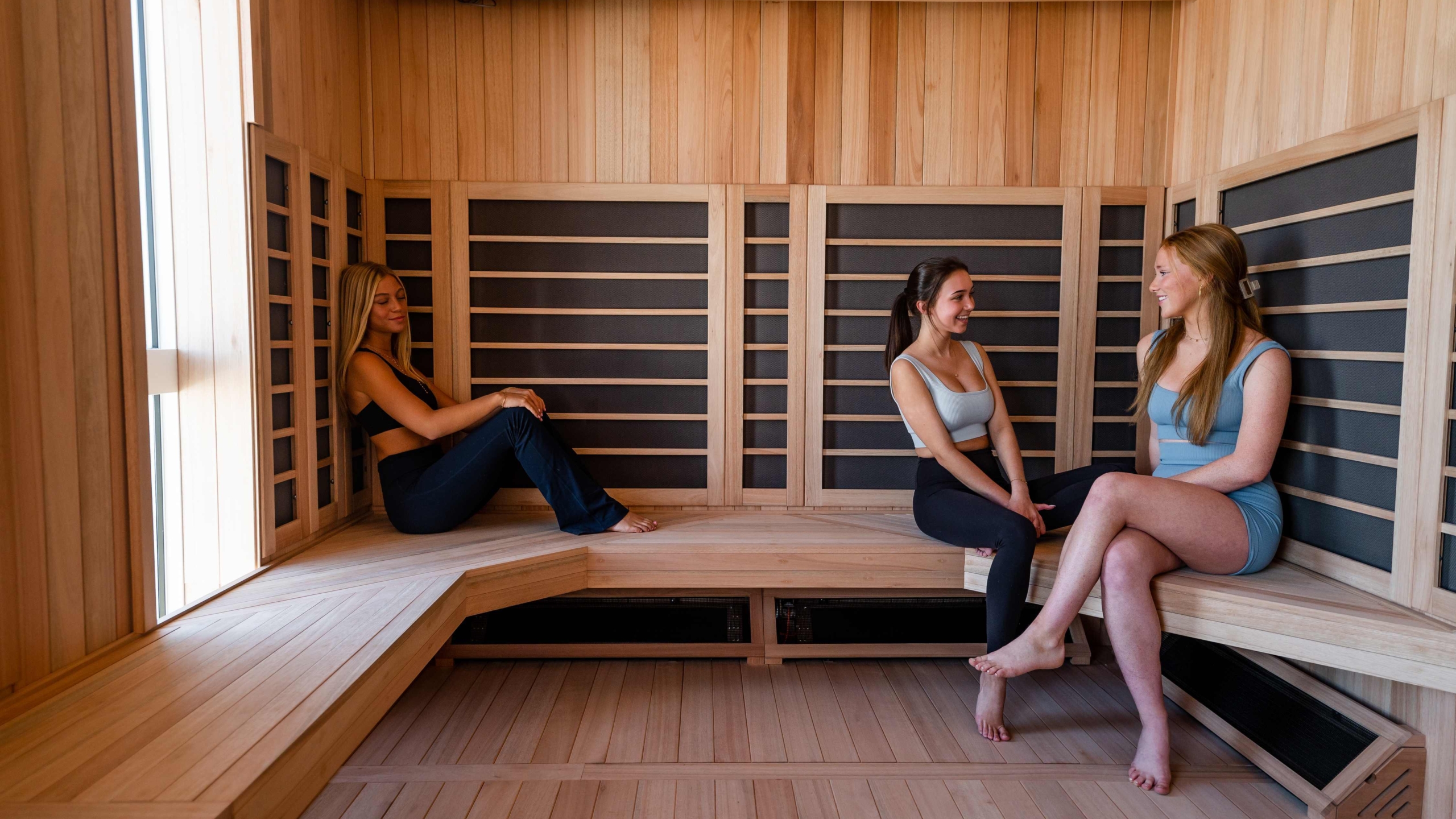 Three women sit and chat together in a modern wooden Sweetwater sauna, two on the right and one on the left, all relaxed and casually dressed in fitness wear. Sunlight enters from a window on the left.