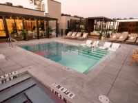 A modern Sweetwater rooftop pool area at sunset features poolside lounge chairs, a hot tub, potted plants, and a glass-walled indoor lounge with warm lighting. The space is clean and inviting.