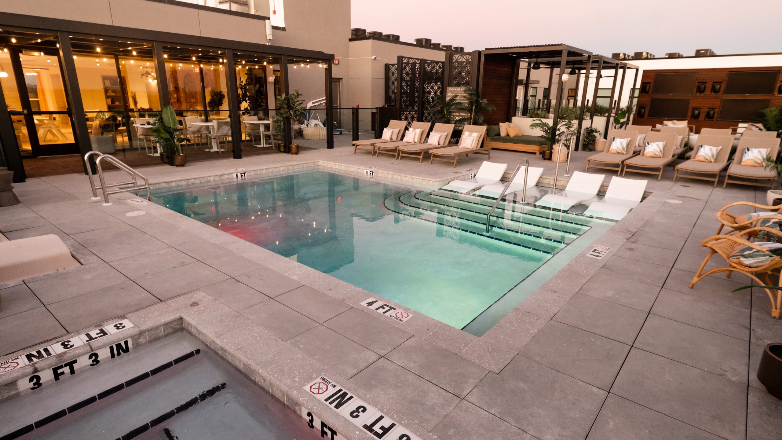 A modern Sweetwater rooftop pool area at sunset features poolside lounge chairs, a hot tub, potted plants, and a glass-walled indoor lounge with warm lighting. The space is clean and inviting.