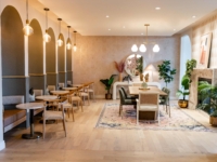 A modern, stylish cafe with arched wall alcoves, small round tables with chairs on the left, a large rug and dining table in the center, warm lighting, indoor plants, and contemporary Sweetwater-inspired decor.