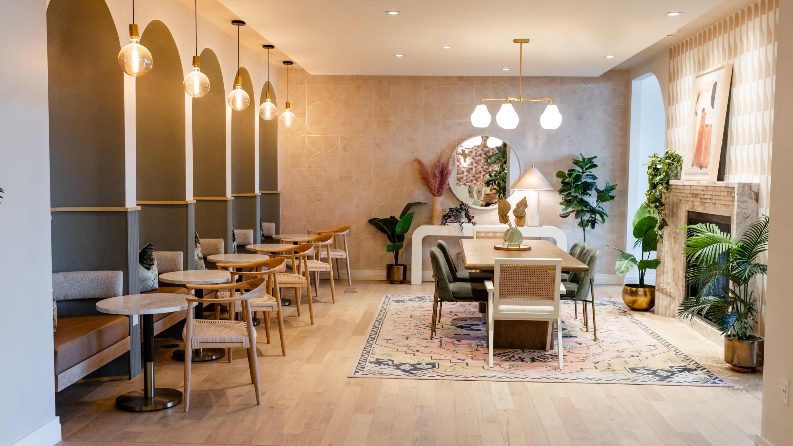A modern, stylish cafe with arched wall alcoves, small round tables with chairs on the left, a large rug and dining table in the center, warm lighting, indoor plants, and contemporary Sweetwater-inspired decor.