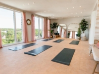 A bright yoga studio at Sweetwater features large windows, pink curtains, black yoga mats, mirrored walls, potted plants, and a cozy seating area with cushions.