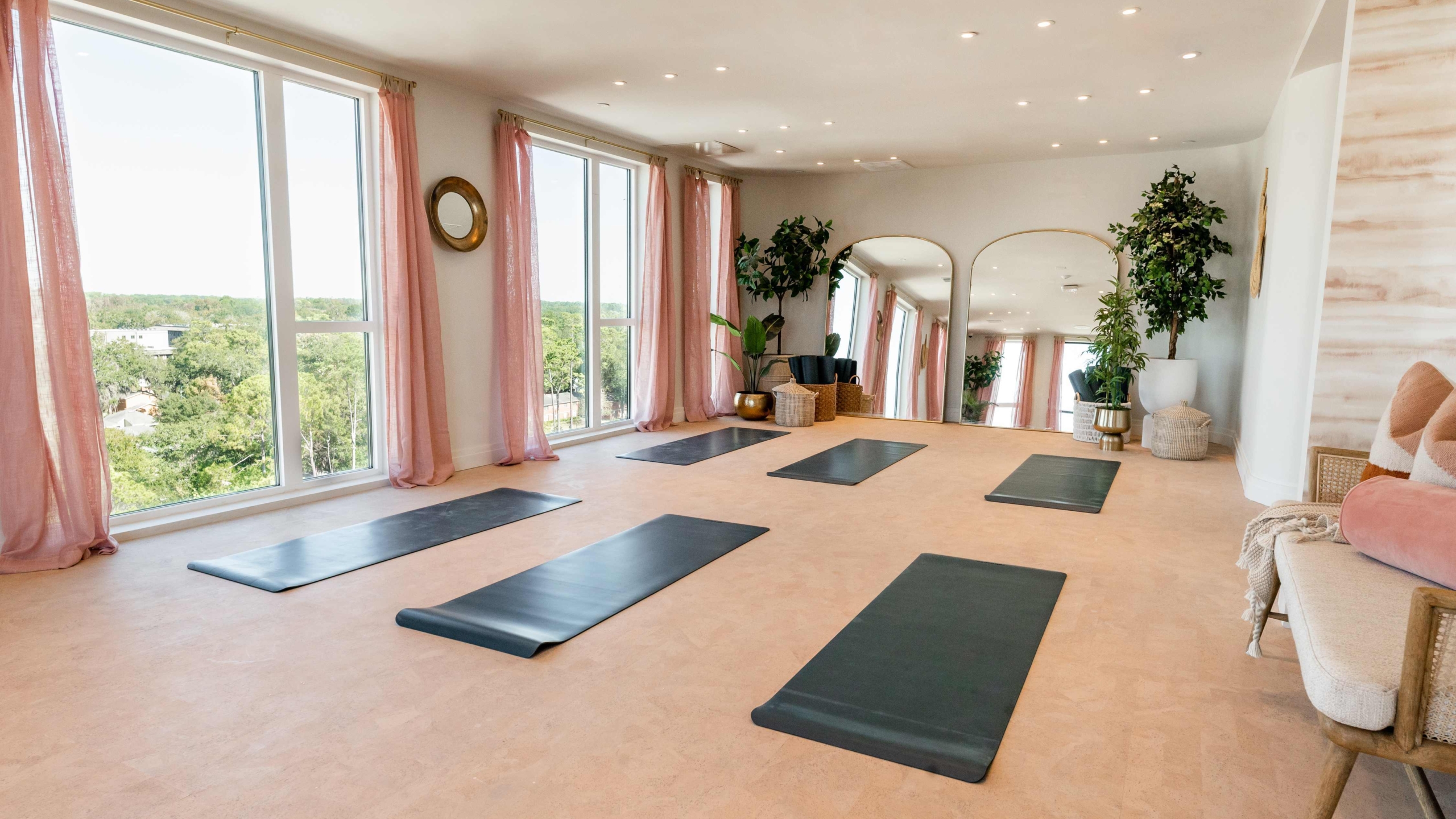 A bright yoga studio at Sweetwater features large windows, pink curtains, black yoga mats, mirrored walls, potted plants, and a cozy seating area with cushions.