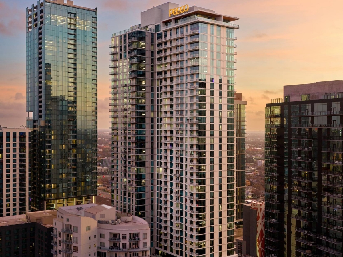 High-rise buildings in a cityscape at sunset, with the tallest tower displaying the sign PMG. The sky glows with warm pink and orange hues as city lights and Paseo below come alive for the evening.