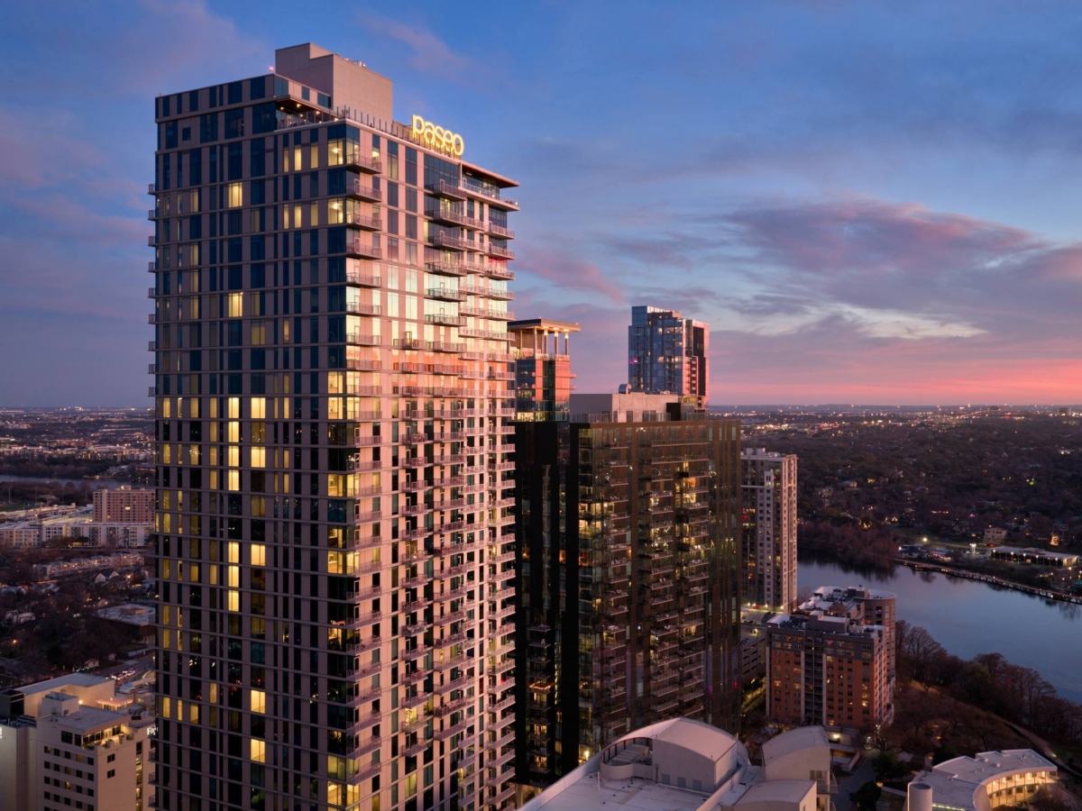 A tall modern apartment building with the sign “Paseo” on top stands against a sunset sky, surrounded by other high-rise buildings and overlooking a river dotted with trees and sparkling city lights in the distance.