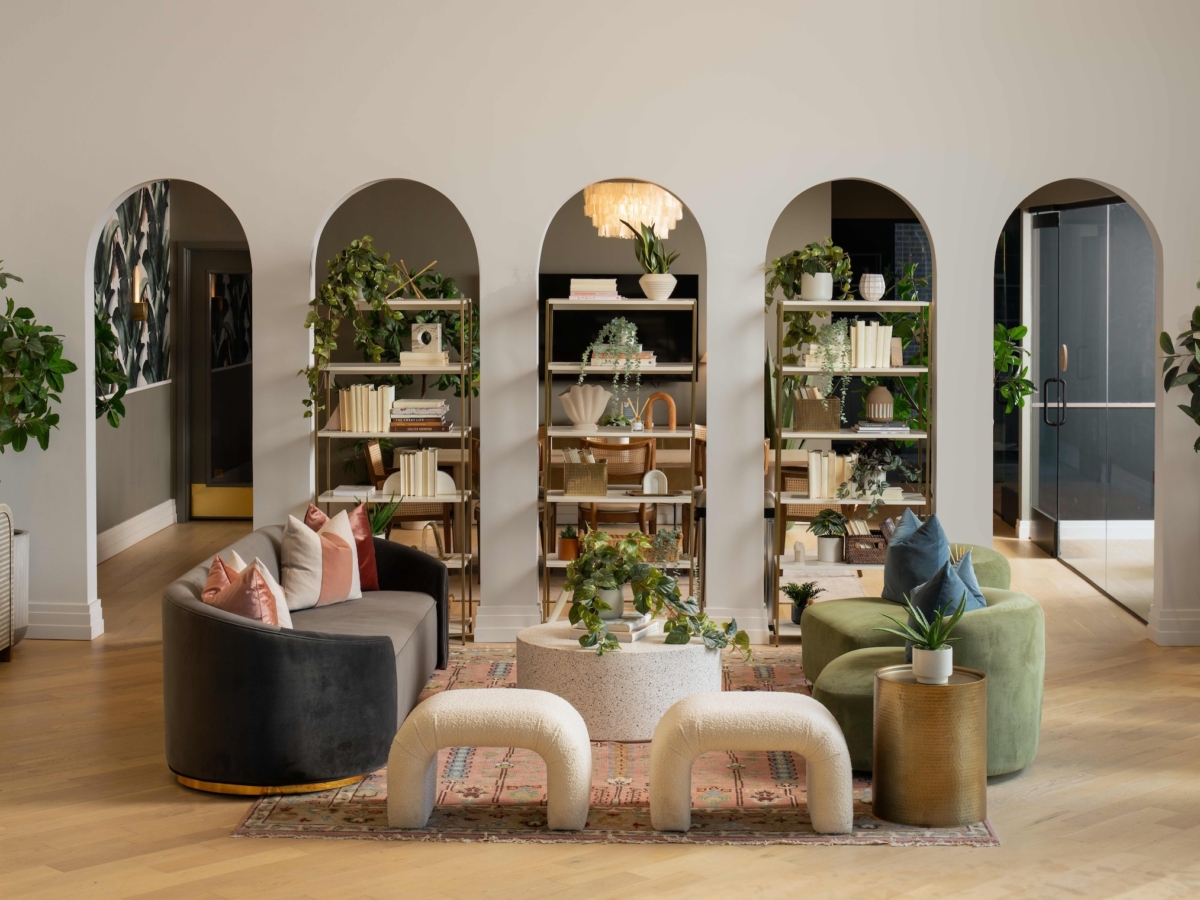 Modern Sweetwater living room with curved sofas, upholstered stools, a round coffee table, and decorative plants. Arched shelves filled with books and greenery are set against a light wall and wooden floor.