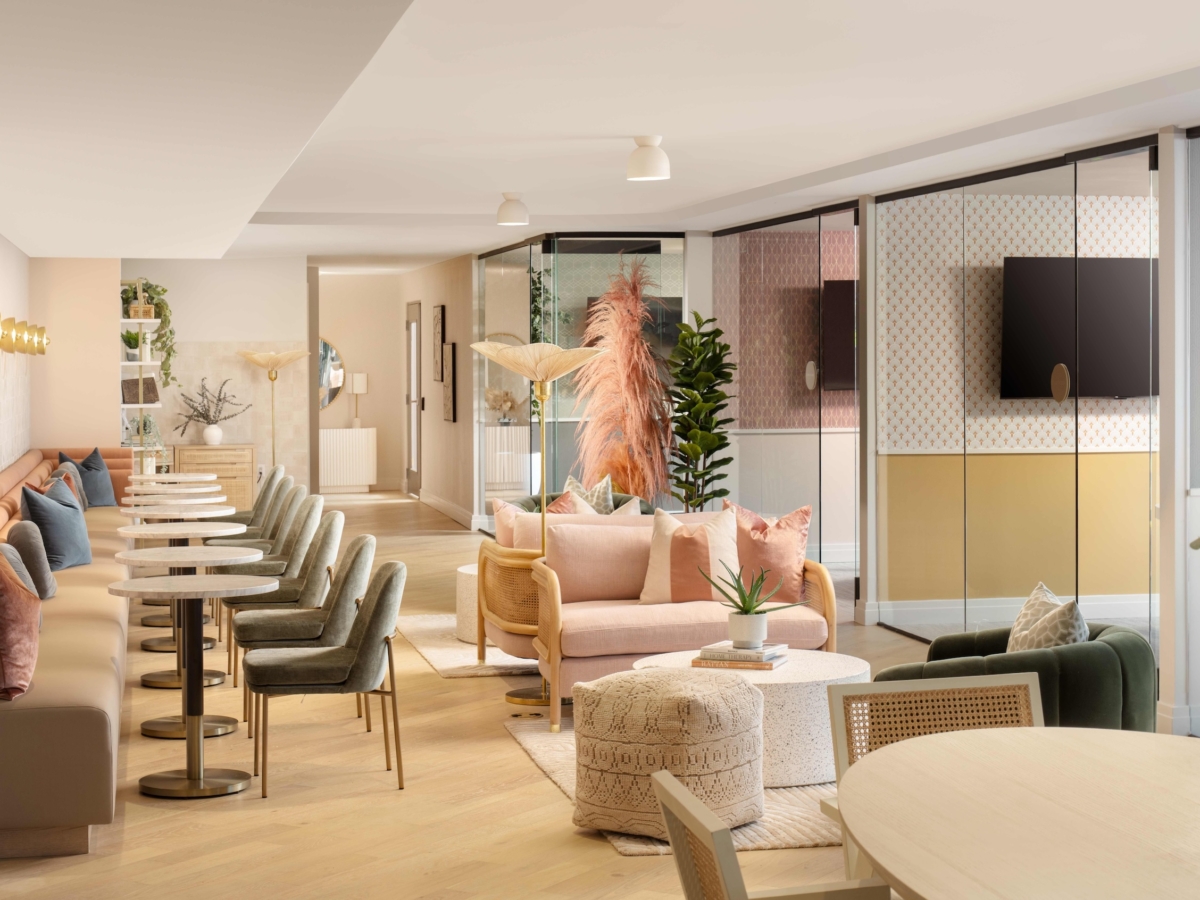 Modern lounge with pastel furnishings, round tables, upholstered chairs, a tan sofa, potted plants, and decorative lighting. Glass walls divide the space while natural light brings a warm Sweetwater vibe to the inviting atmosphere.