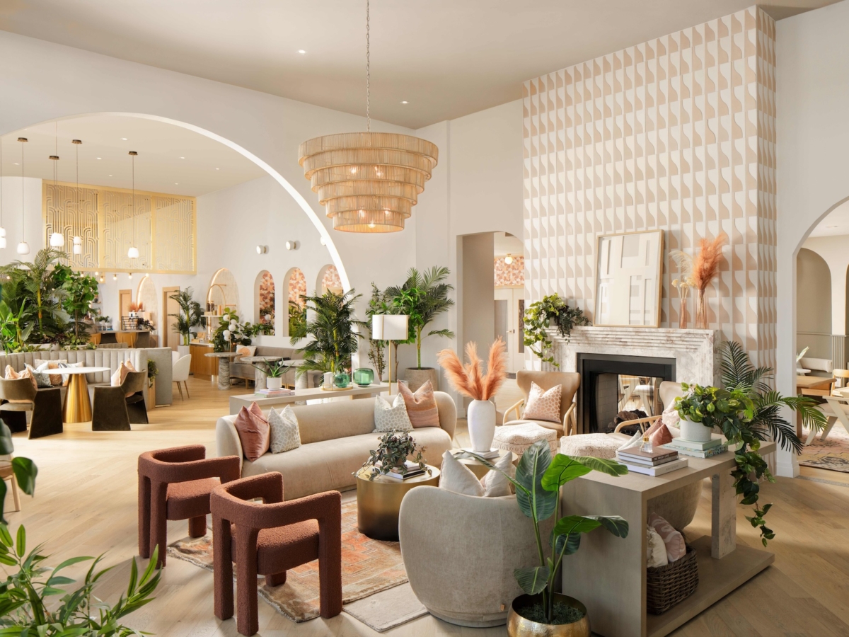 A spacious, modern lounge with neutral tones, light wood floors, leafy plants, and cozy seating. Featuring a decorative fireplace, a large chandelier, stylish Sweetwater-inspired decor, and geometric patterns on the walls.