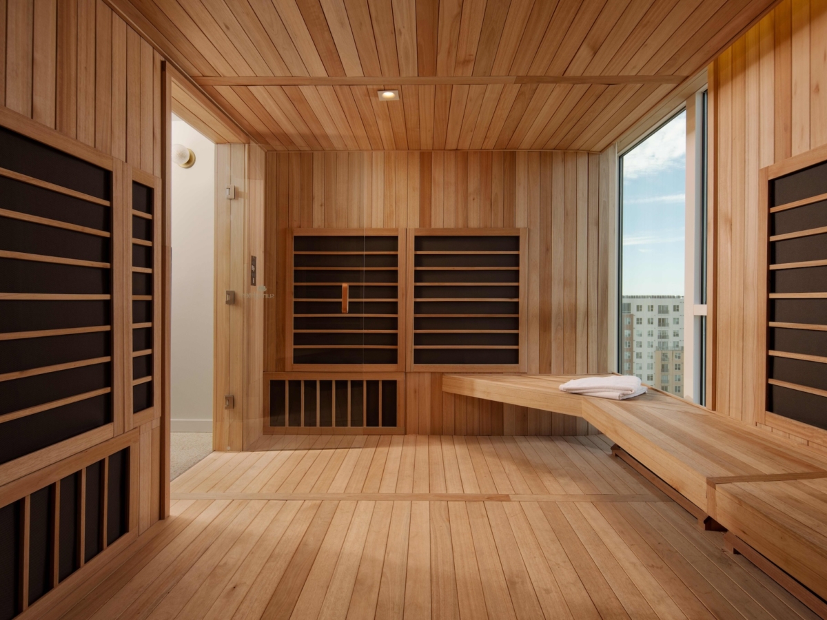 A modern Sweetwater sauna room with wooden walls, ceiling, and floor features black infrared panels, a built-in wooden bench with folded towels, a glass window, and a door leading outside. Daylight streams in through the window.