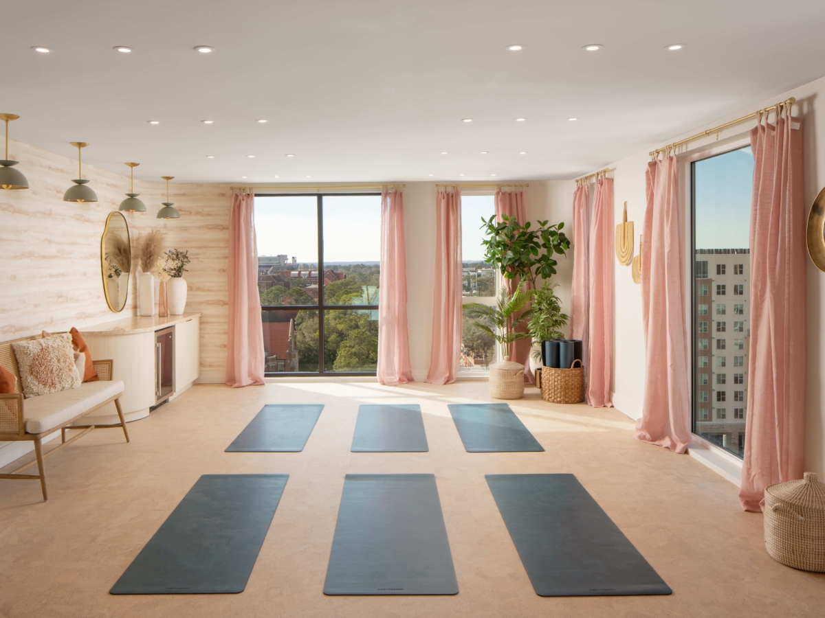 A bright yoga studio with six black mats on the floor, pink curtains, large windows with Sweetwater city views, plants, and gold accents like mirrors and lighting. The room feels modern, airy, and inviting.