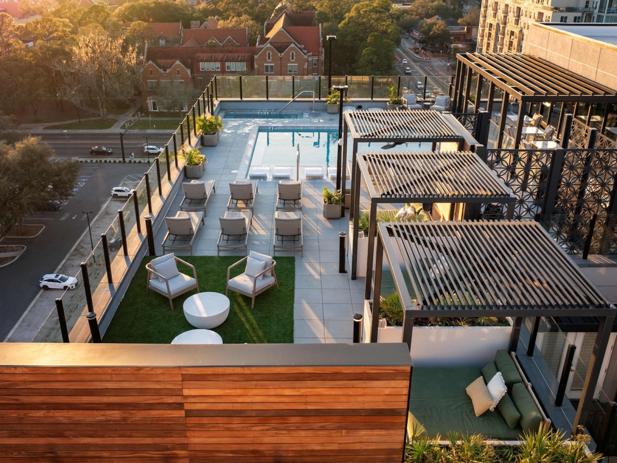 A modern rooftop lounge at Sweetwater features sun loungers by a pool, shaded seating areas with chairs and tables, and stunning city views at sunset. Trees and buildings complete the vibrant backdrop.