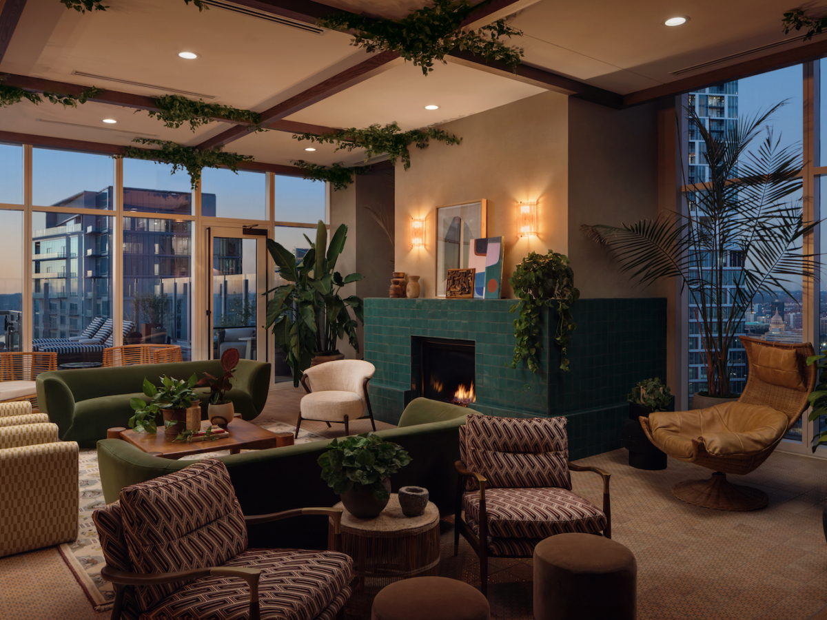 A stylish Paseo lounge with modern furniture, green tiled fireplace, plants, large windows, and city views at dusk. Cozy seating includes sofas, armchairs, and a tan lounge chair, with greenery and warm lighting throughout.
