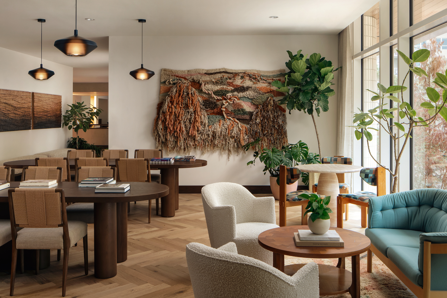 A stylish, modern lounge with wooden tables, patterned chairs, plush armchairs, large indoor plants, and textured wall art. Sunlight streams through floor-to-ceiling windows, creating a warm and inviting Paseo-inspired atmosphere.