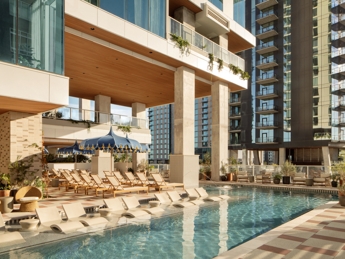 Modern outdoor pool area at Paseo features lounge chairs partially submerged in water, surrounded by high-rise buildings and shaded seating under a blue and white canopy. Tall glass windows and lush greenery complete the inviting space.