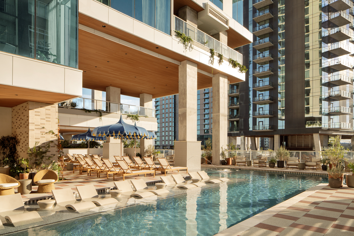 Modern outdoor pool area at Paseo features lounge chairs partially submerged in water, surrounded by high-rise buildings and shaded seating under a blue and white canopy. Tall glass windows and lush greenery complete the inviting space.