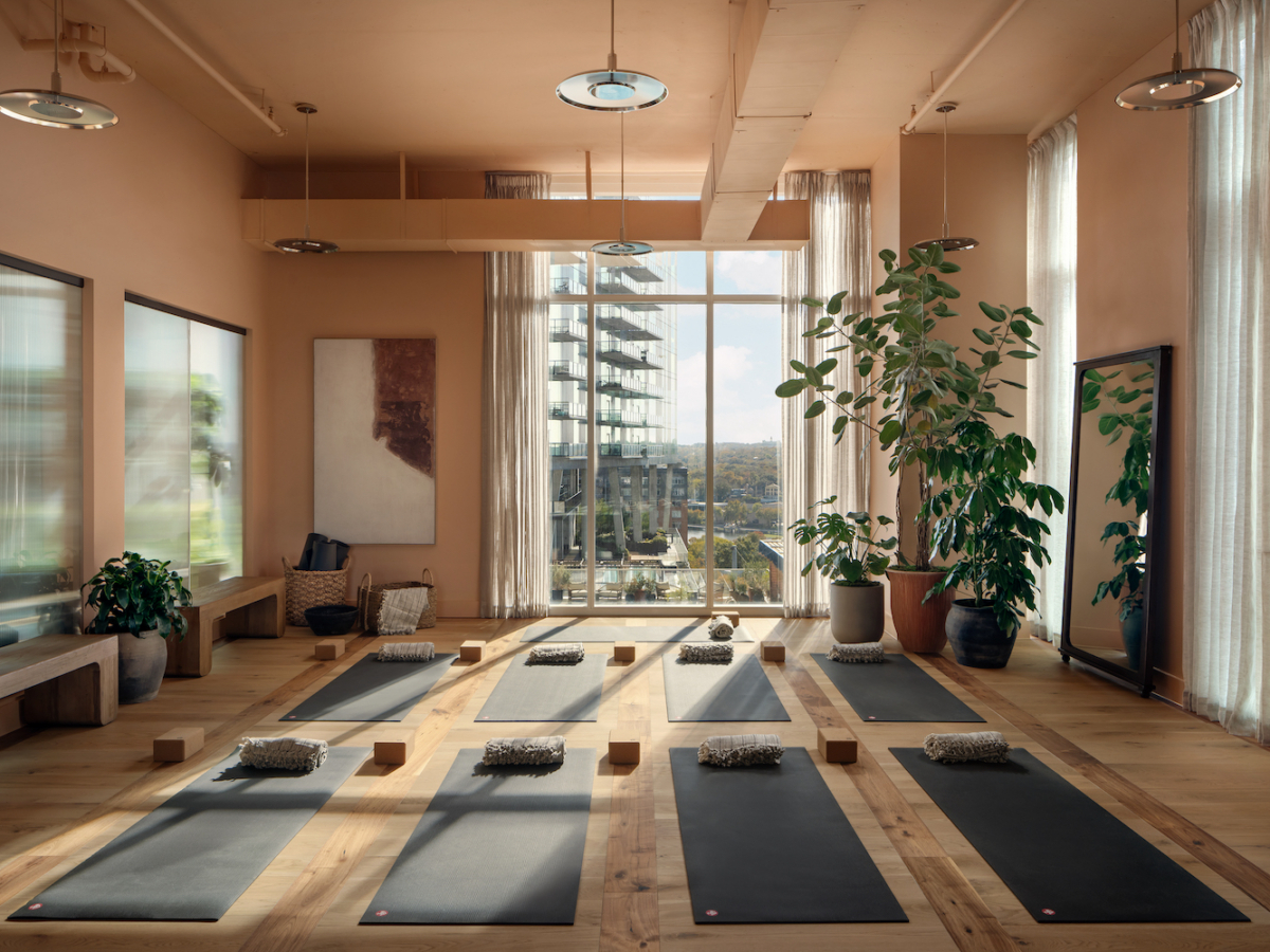 A spacious, sunlit yoga studio on Paseo with large windows, wooden floors, mats, cushions, and blocks neatly arranged. Potted plants and mirrors line the room, while a city view is visible through the windows.