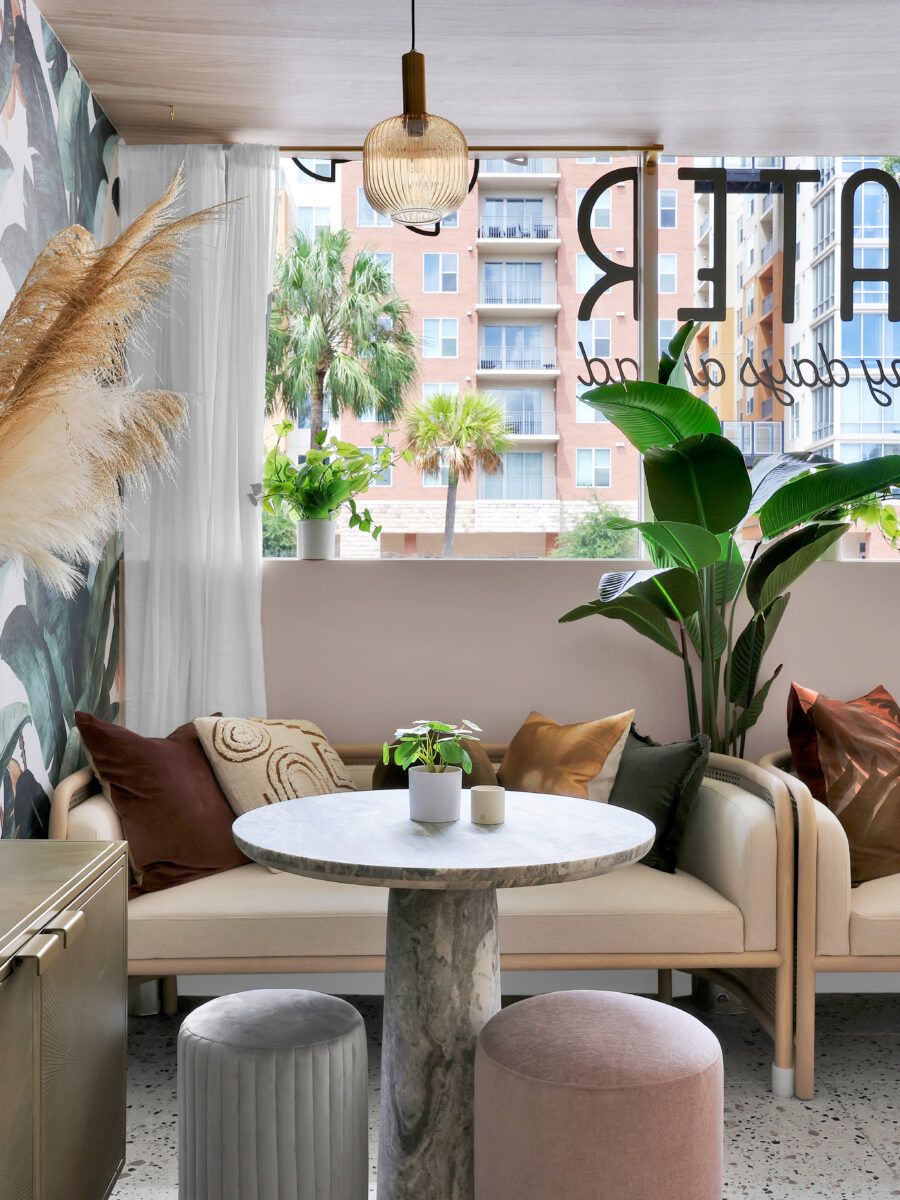 Modern café interior with tropical wallpaper, plants, round marble tables, plush seating with cushions, and soft natural light from large windows. Urban buildings and palm trees frame the view outside this inviting Addy space.