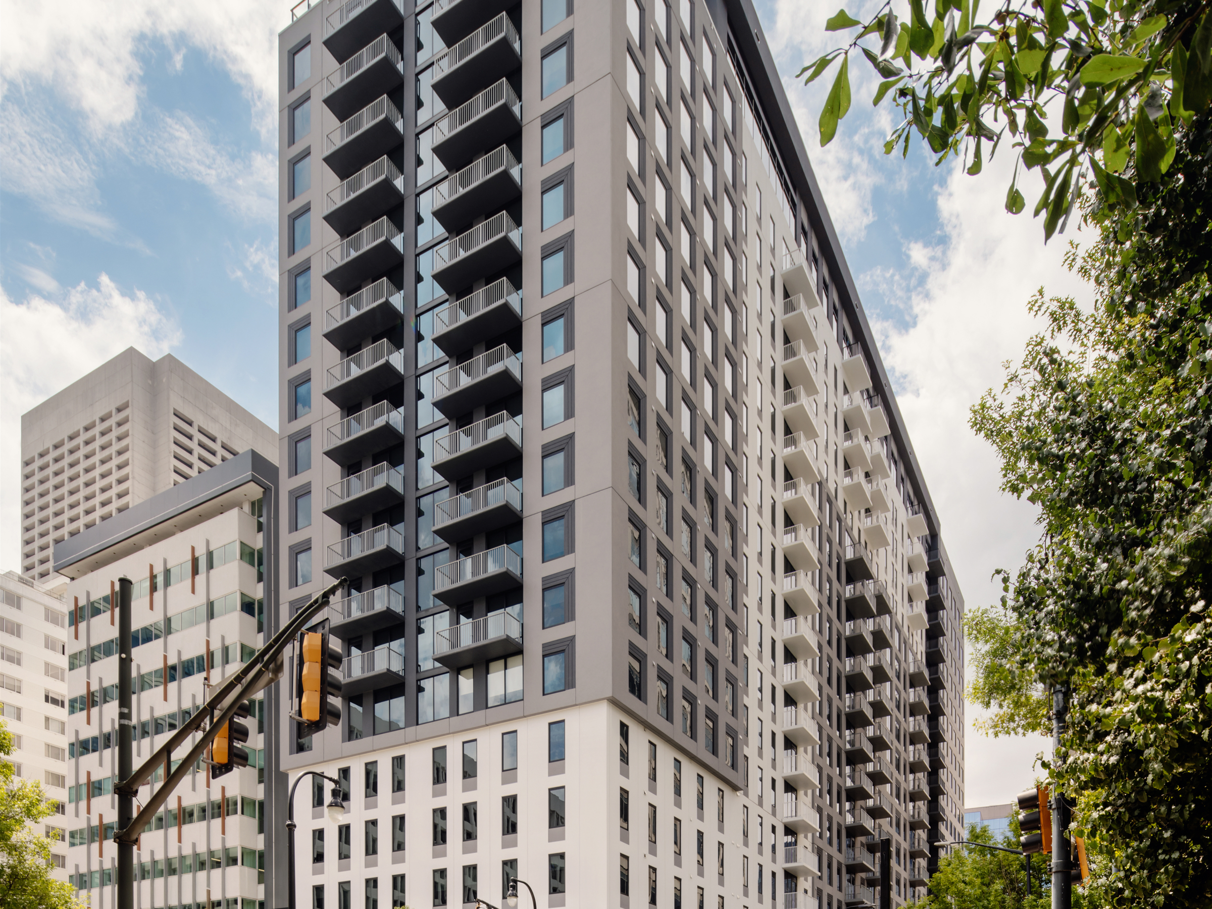 A modern high-rise apartment building, like Rambler Atlanta, stands on a city corner with people crossing the street, surrounded by other tall buildings, trees, and traffic lights under a partly cloudy sky.