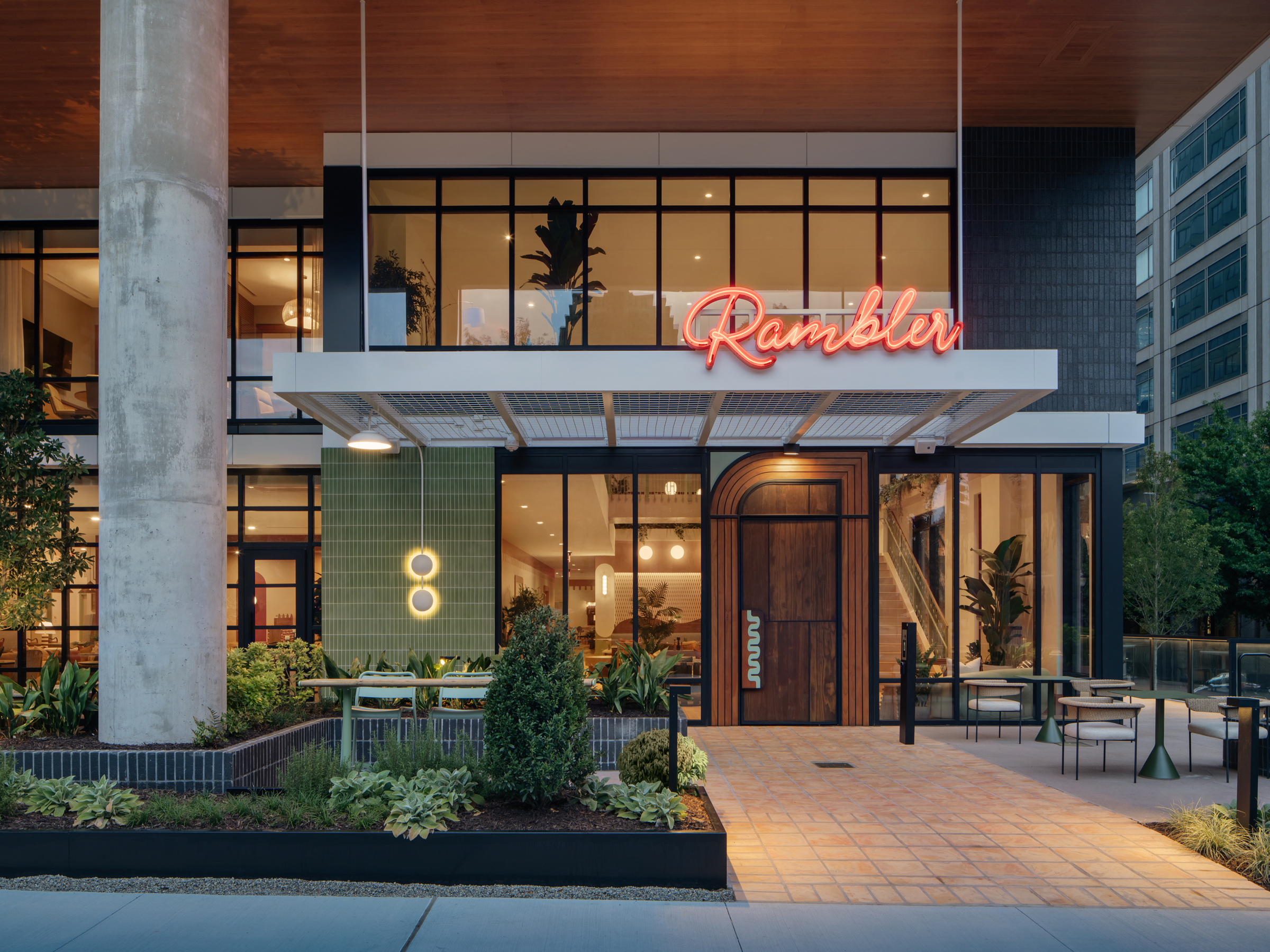 Modern building entrance with large windows, greenery, patio seating, and a neon Rambler Atlanta sign above the door. Warm lighting and stylish decor are visible inside through the glass.
