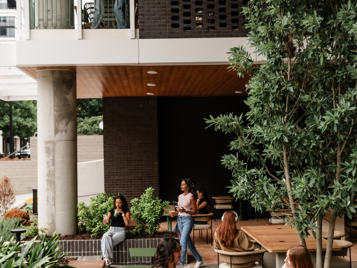 A modern outdoor patio scene at Rambler Atlanta with people socializing. Two women sit at a wooden table, while others mingle near planters and a large tree beneath the cafe’s elevated balcony.