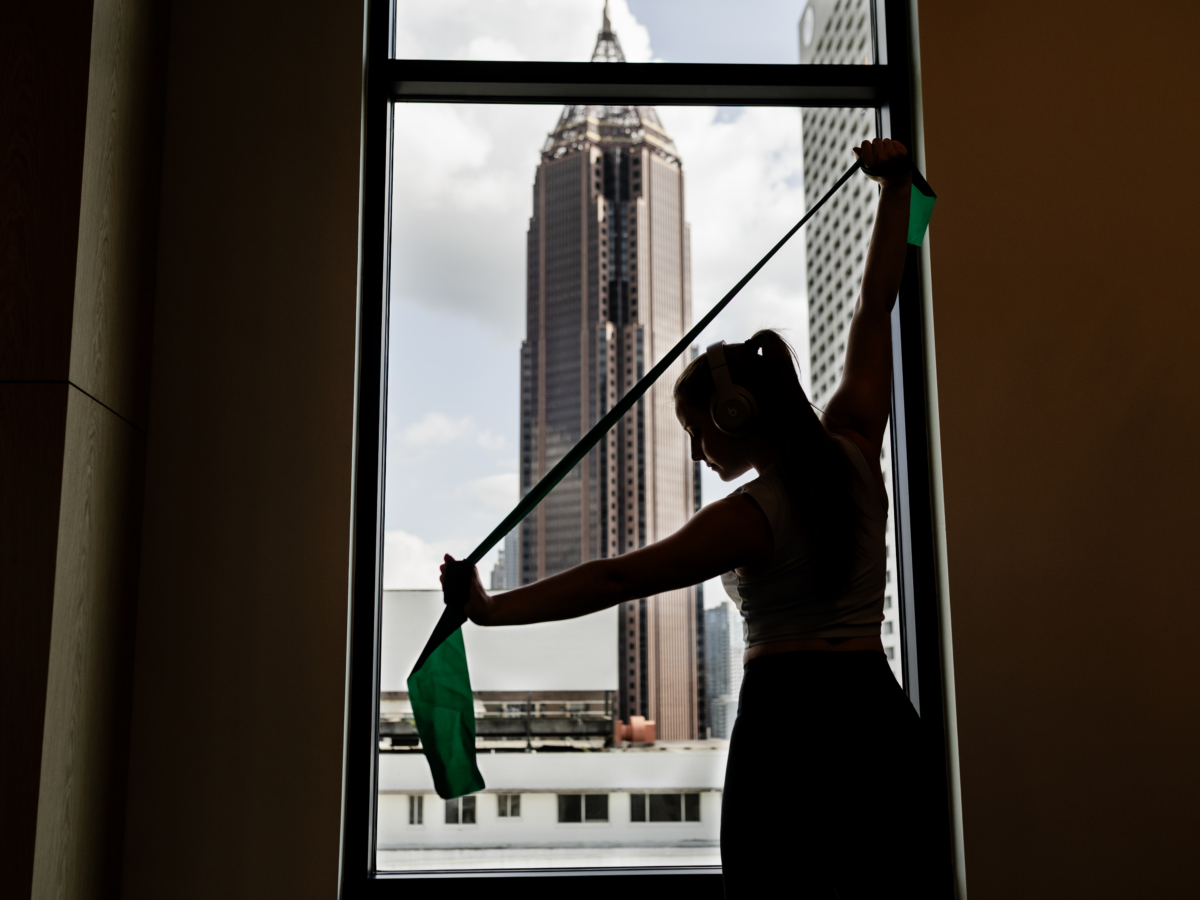 A woman silhouetted against a large window at Rambler Atlanta stretches a resistance band, with a tall city building visible outside under a partly cloudy sky.