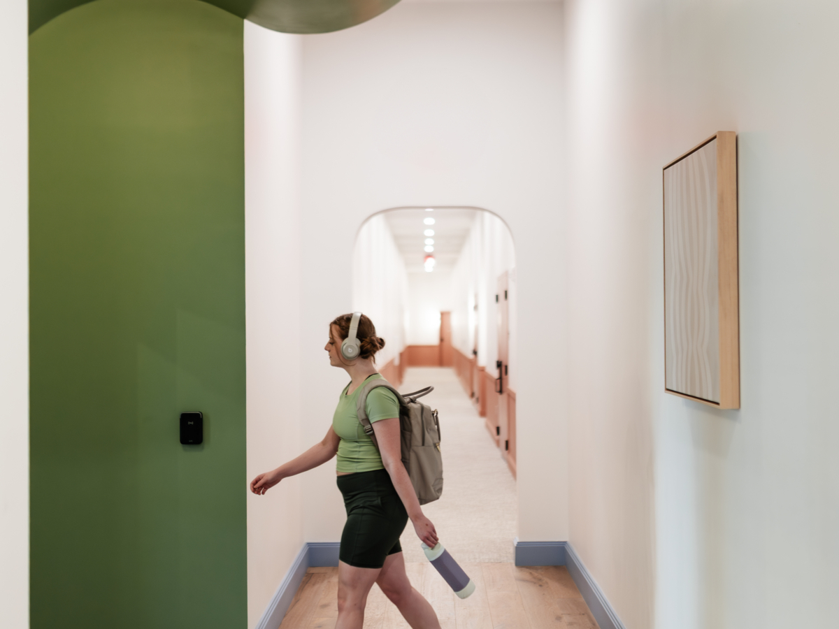 A person with a backpack and yoga mat walks through a hallway at Rambler Atlanta, featuring curved green and white walls, patterned tile flooring, and soft lighting.