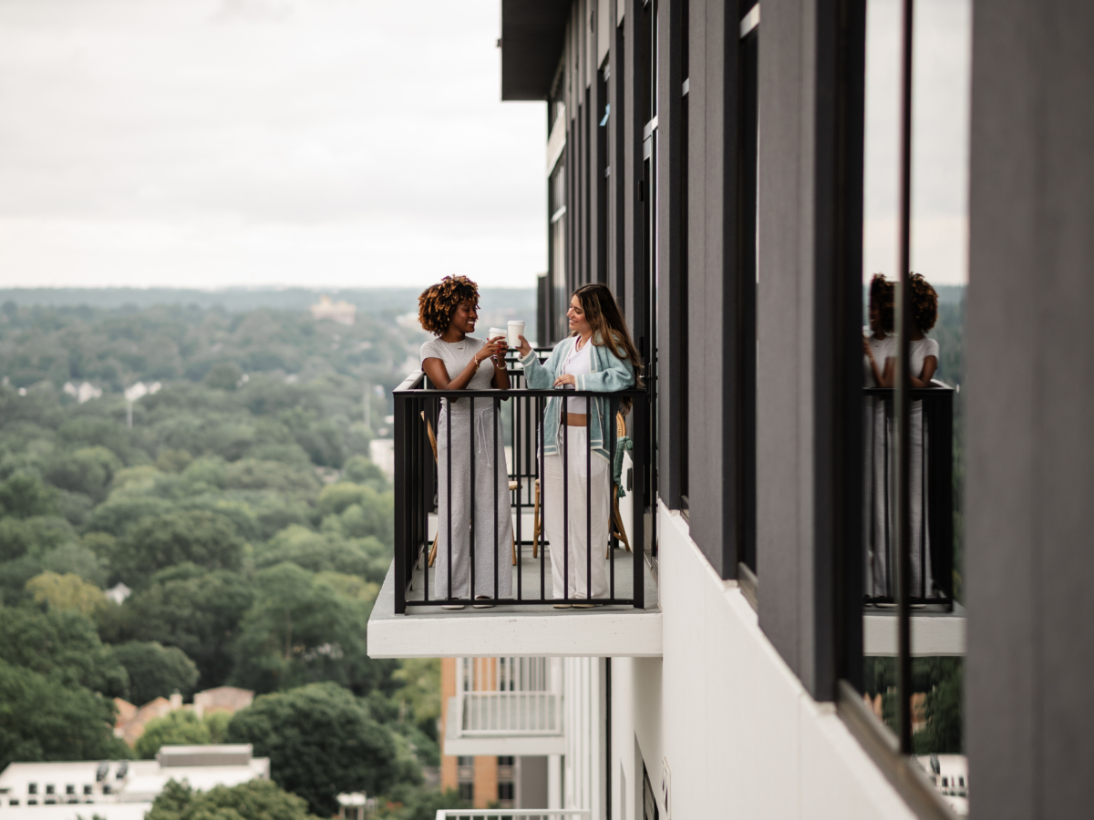 Two women stand on a high-rise balcony at Rambler Atlanta, smiling and clinking glasses, surrounded by a scenic view of trees and buildings in the distance.