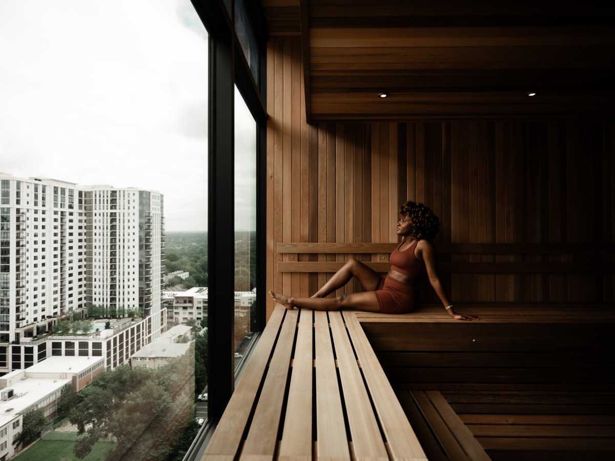 A woman sits on a wooden bench in a modern sauna at Rambler Atlanta, gazing out large windows at tall buildings and the city skyline during the day.