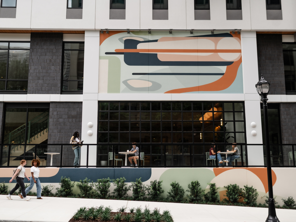 People walk and sit outside Rambler Atlanta, a modern building with large windows and a colorful abstract mural. Some pass by while others relax at tables on the elevated walkway lined with plants below.