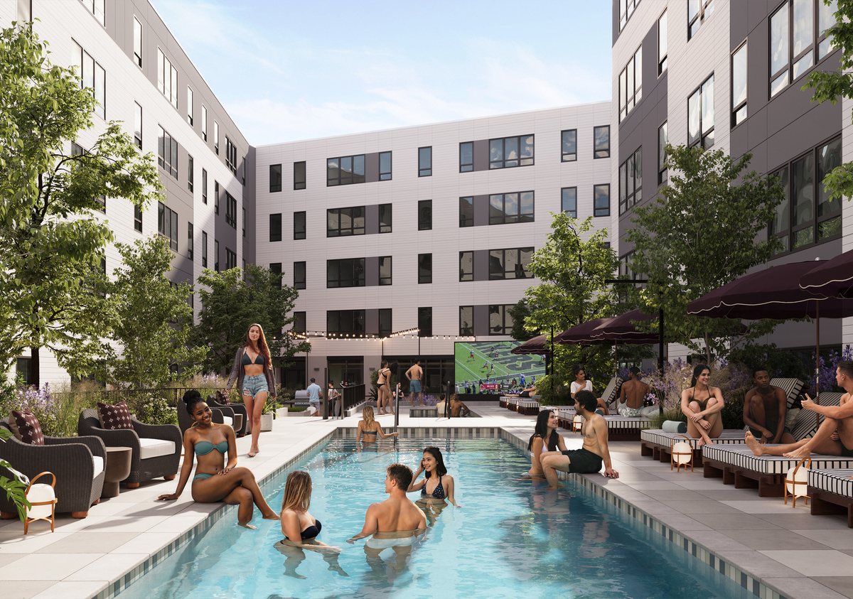 People relax and socialize around the outdoor pool at Rambler Riverfront's modern apartment courtyard. Some lounge on chairs under umbrellas, while others wade in the water. Trees and plants decorate the area for a lively, welcoming atmosphere.