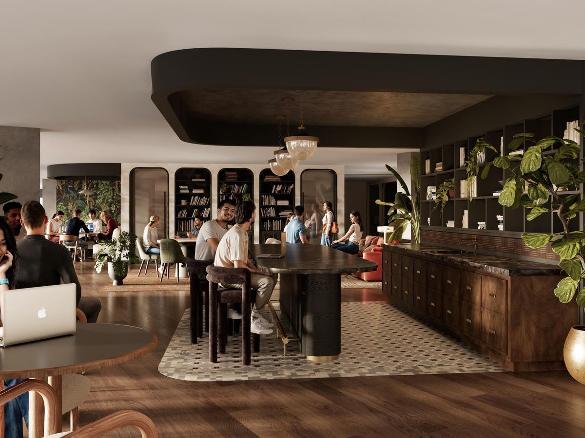 A spacious, modern co-working space at Rambler Riverfront with people working and socializing. There are wooden floors, large plants, bookshelves, and a central island with stools. A woman smiles while using a laptop at a round table in the foreground.