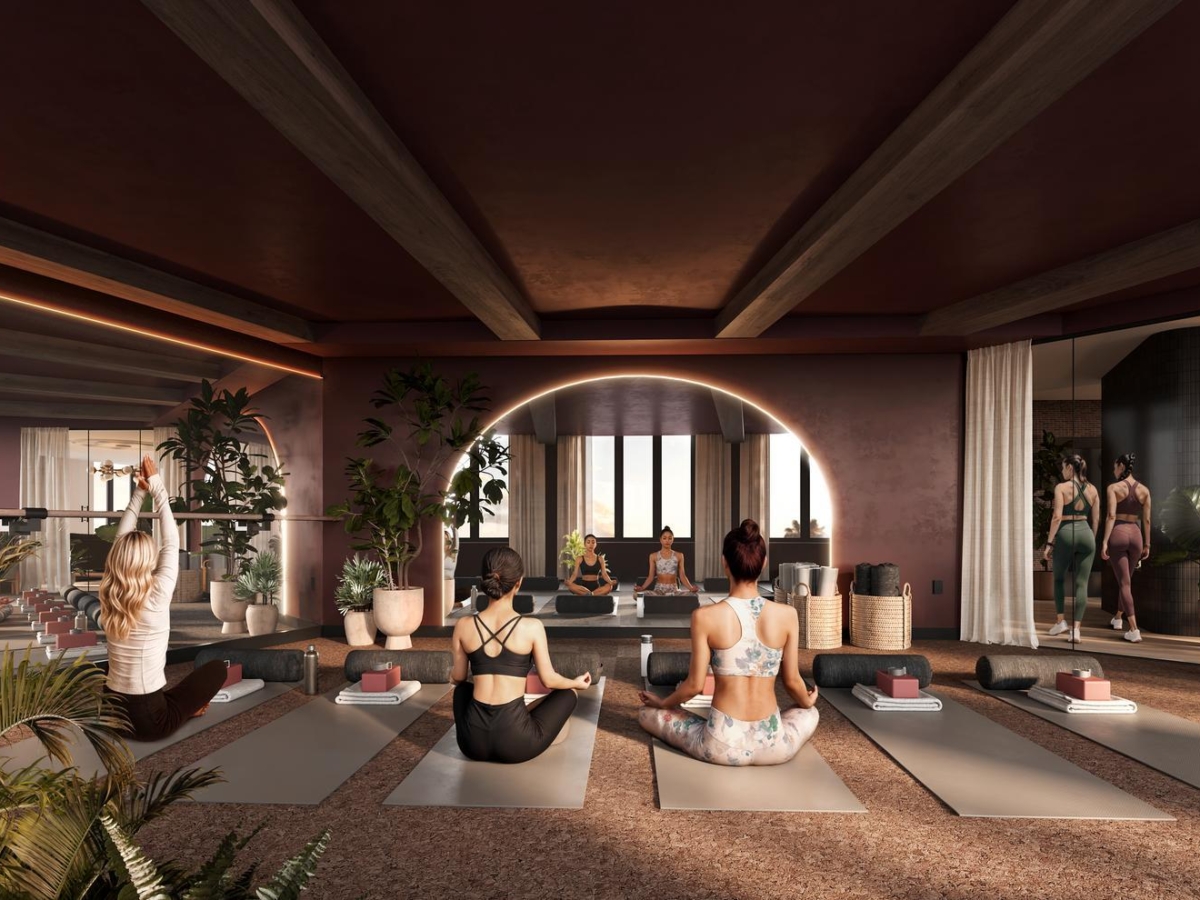 A group of people practice yoga in a modern, stylish Rambler Riverfront studio with large mirrors, potted plants, and soft lighting. Three women sit on yoga mats in the foreground, facing an instructor at the front of the room.