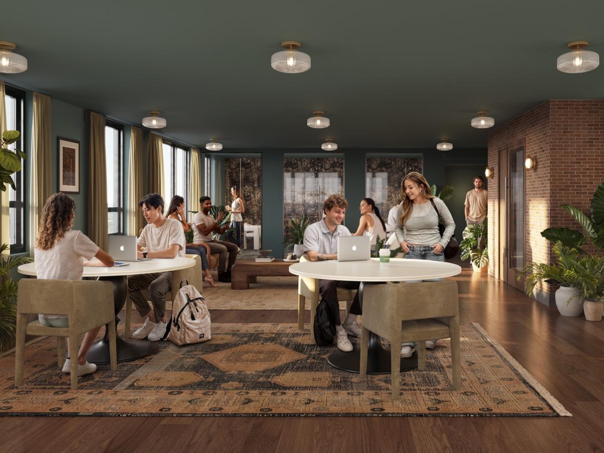 Modern lounge at Rambler Riverfront with large windows, plants, and rugs. Several young adults sit and stand around tables, talking, using laptops, and writing. Warm lighting and cozy décor create a collaborative workspace.