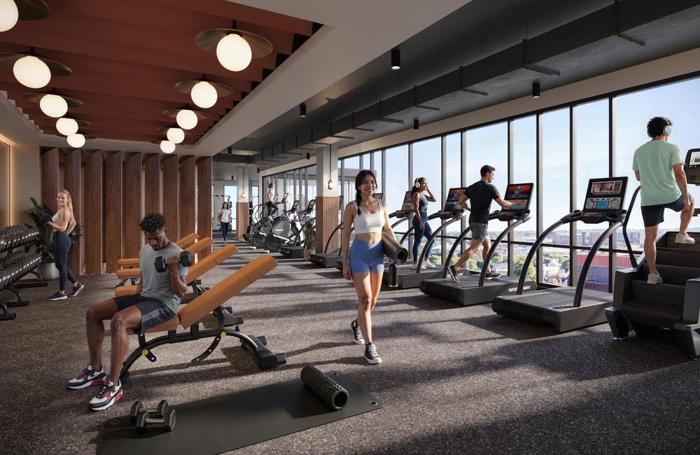 People working out in the modern Rambler Ann Arbor gym with large windows. Some use treadmills and stair machines, while others lift weights or stretch on the floor. Bright, natural light fills the spacious room.