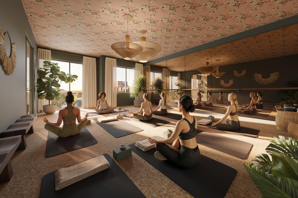 A group of people practice yoga on mats in the spacious, sunlit Rambler Ann Arbor studio with large windows, plants, and decorative lighting. Seated in meditation poses facing a mirrored wall, they enjoy a calm and peaceful atmosphere.
