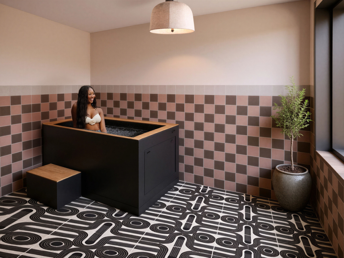 A woman in a white bikini relaxes in a modern black plunge pool at Rambler Ann Arbor, surrounded by pink and brown walls, geometric black-and-white floor tiles, a potted plant, and natural light streaming through a large window.