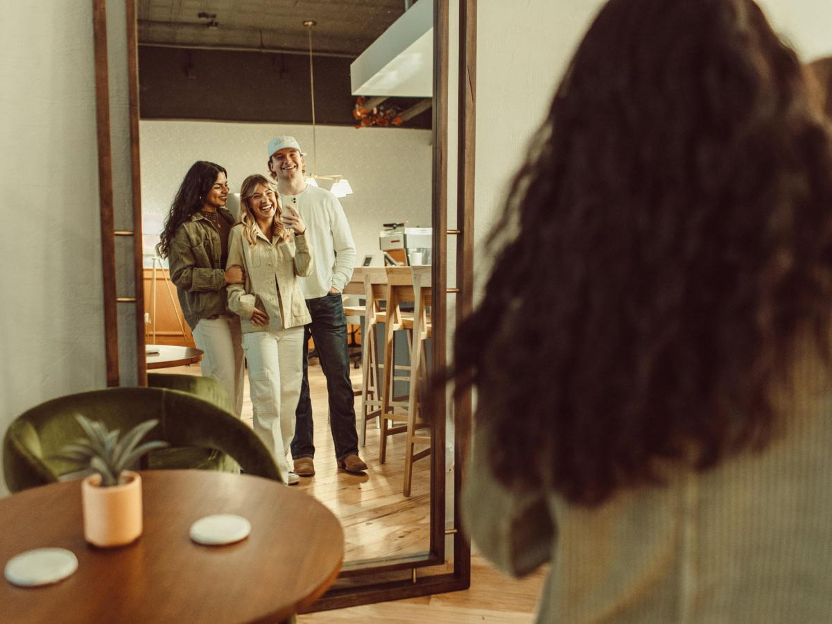 Three people smile and pose together in front of a large mirror in a cozy LV Collective Rambler setting, while another person with long hair captures their photo from behind. A small round table with a potted plant sits in the foreground.
