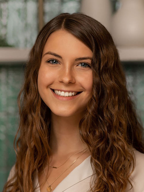 A young woman with long wavy brown hair and blue eyes smiles at the camera. She is wearing a white top and a layered necklace, standing indoors with a blurred background of shelves and decorative items.