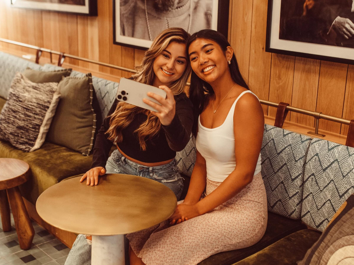 Two young women sit on a green velvet bench in a stylish cafe, smiling and taking a selfie together. They are surrounded by decorative pillows and large black-and-white portraits on the wooden-paneled wall behind them.