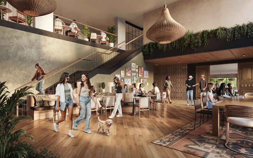 A spacious, modern lounge with wooden floors, large windows, and greenery. People are socializing, working, and relaxing; some sit at tables, others walk or talk, and one woman walks a small dog. A staircase leads to an upper level.