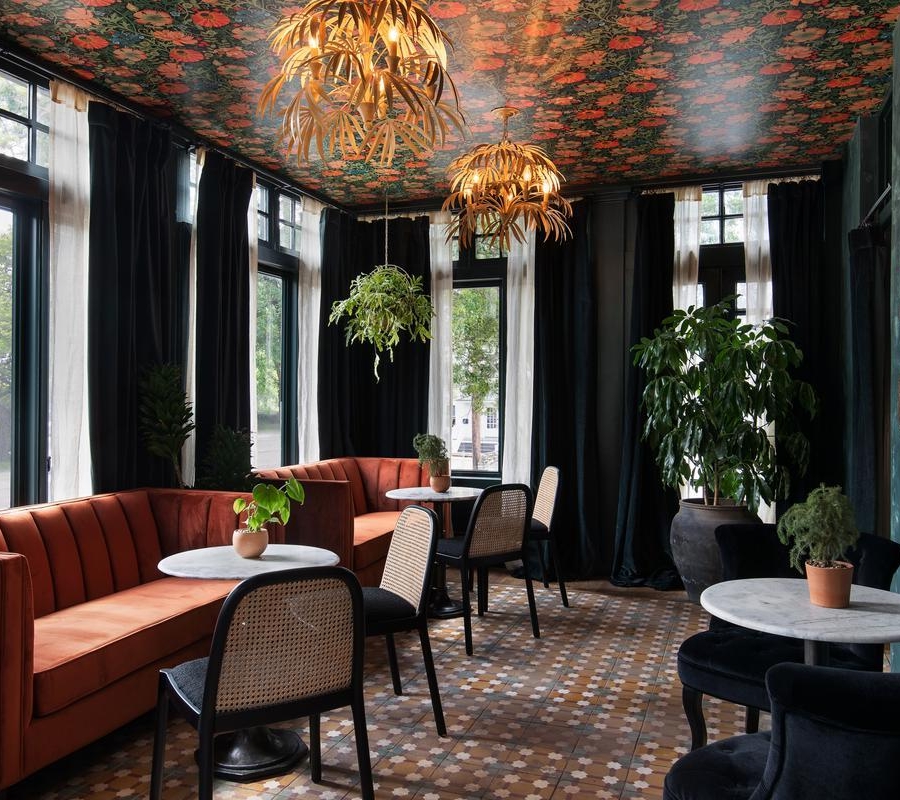 A stylish lounge with orange velvet sofas, round marble tables, and wicker chairs. Potted plants, large windows with black curtains, floral ceiling, and copper wall decor create a cozy, elegant atmosphere.