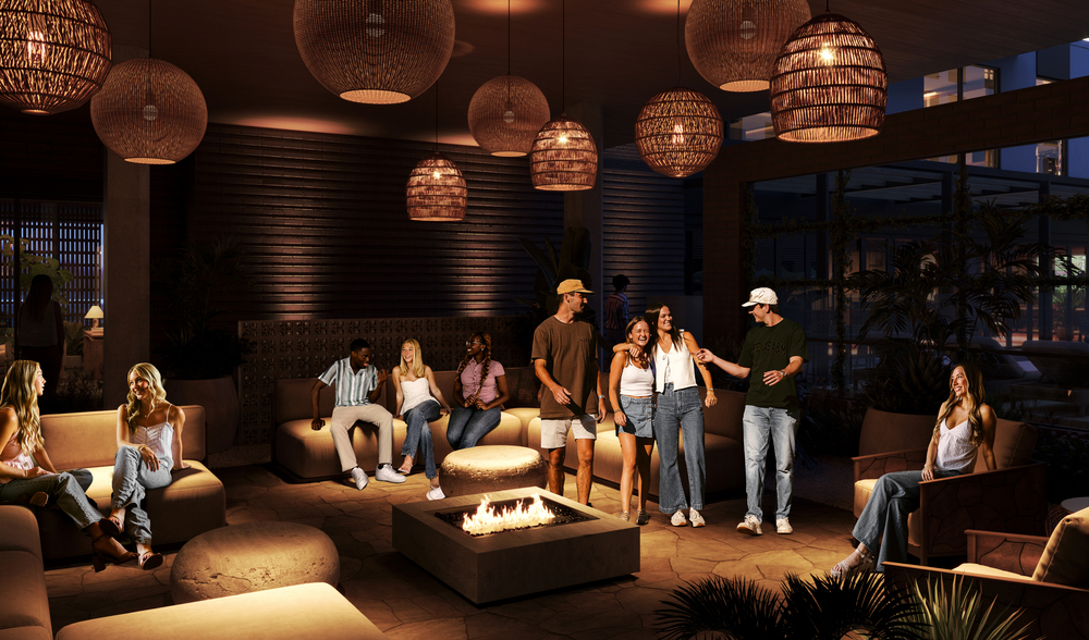 A group of young adults relax and socialize around a fire pit in a stylish, warmly lit lounge with modern decor, woven hanging lamps, and comfortable seating at night.