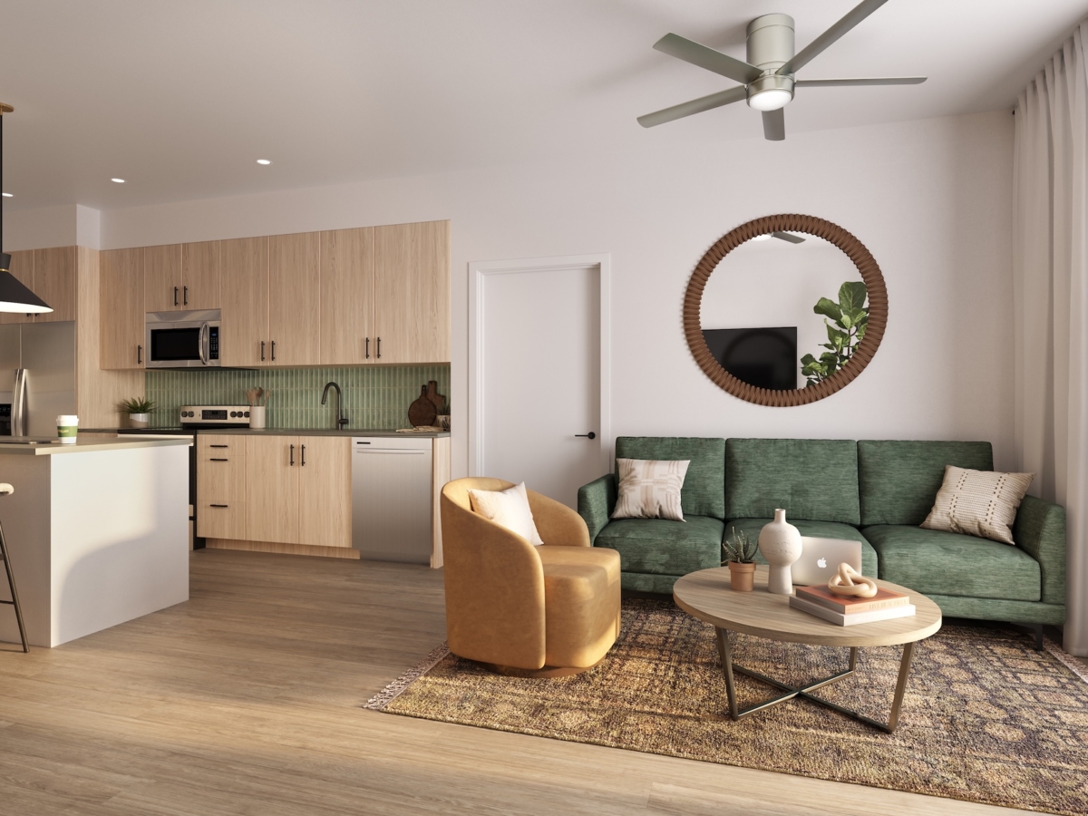 Modern apartment living room with an open kitchen, green sofa, round coffee table, tan armchair, large window, ceiling fan, and wall mirror above the couch. The space is bright with natural light and contemporary decor.