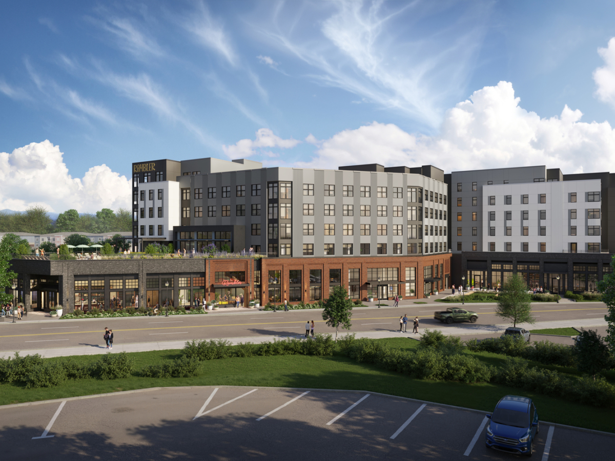 Modern multi-story building with gray and white exterior, ground-floor shops, and large windows, surrounded by greenery, a few parked cars, and people walking along the sidewalk under a partly cloudy sky.
